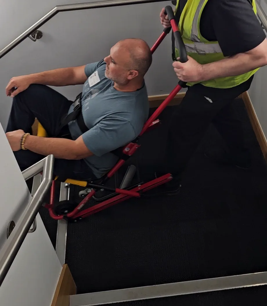 An operator in a hi-vis vest bringing a seated man down a stairwell in a HALO evacuation chair during training