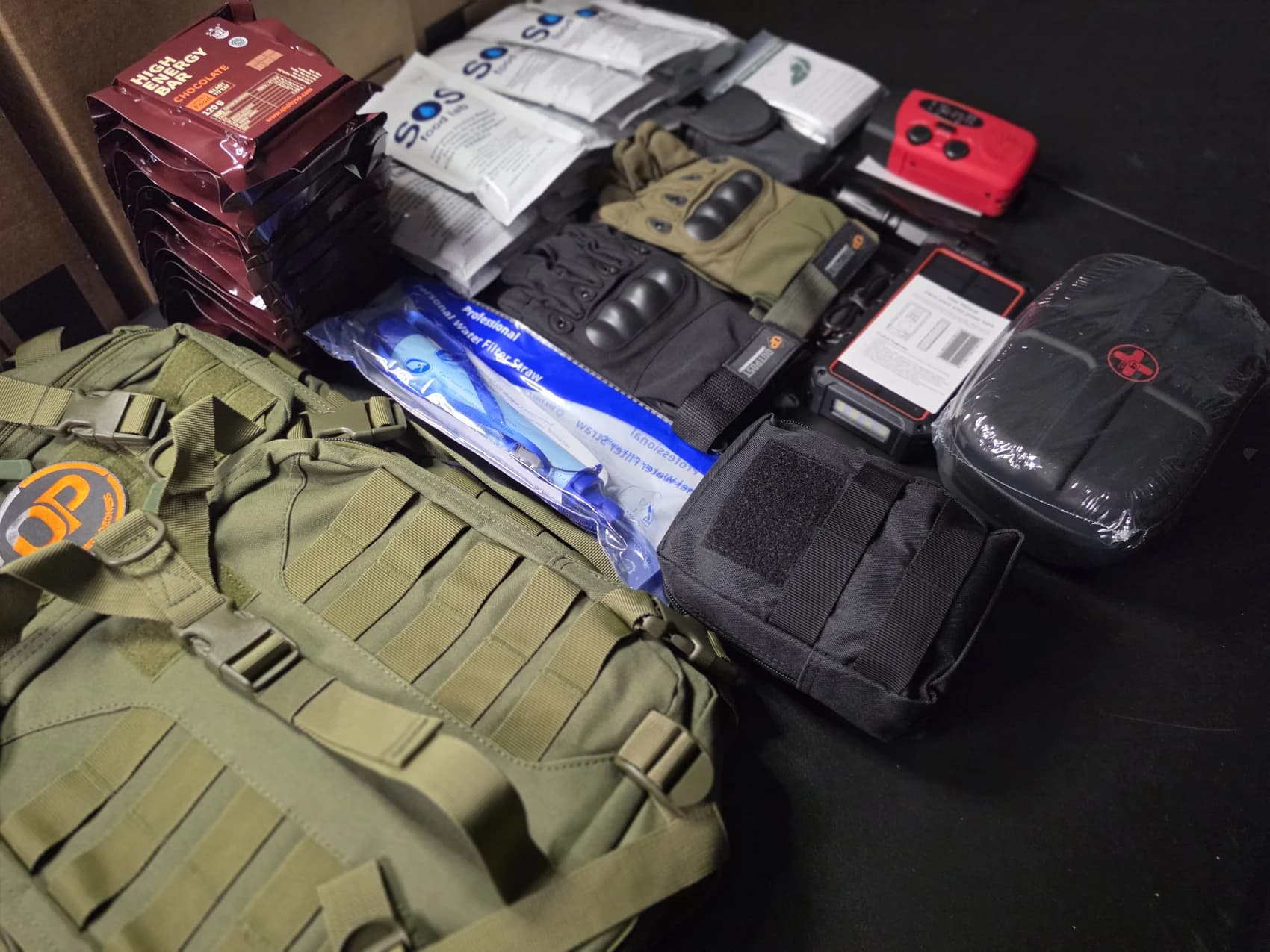 An Outpost go-bag with its contents laid out: ration packs, water pouches, gloves, water filter straw, emergency radio, solar power bank, flashlight and medical kit